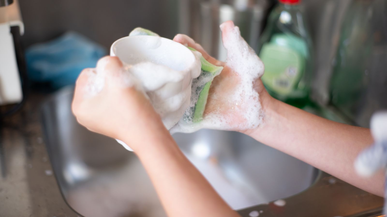 Hands washing an air fryer basket with soap and water in a kitchen sink